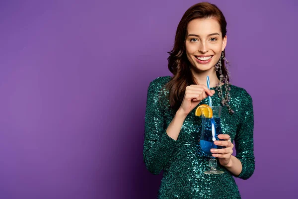 Cheerful woman in party dress holding alcohol cocktail and straw on purple — Stock Photo