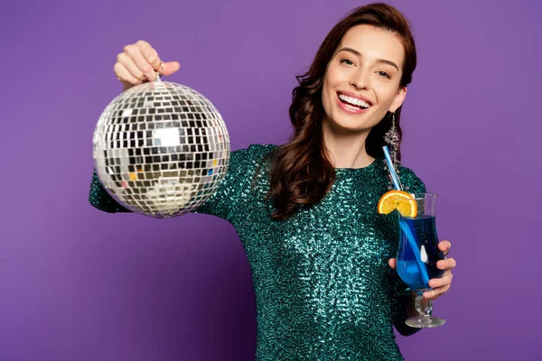 Happy woman in dress holding disco ball and cocktail in glass on purple — Stock Photo