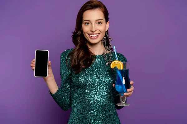 Щаслива молода жінка в сукні тримає коктейль і смартфон з порожнім екраном на фіолетовому — стокове фото