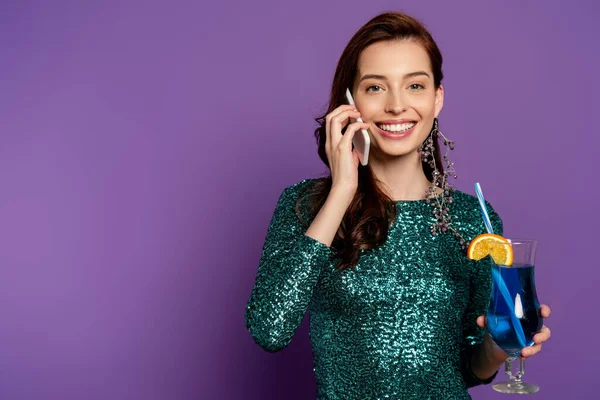 Cheerful young woman in dress holding cocktail and talking on smartphone on purple — Stock Photo