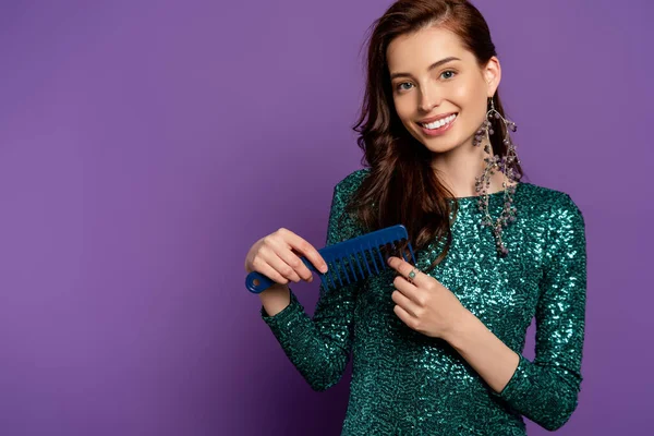 Cheerful woman in dress holding comb and brushing hair on purple — Stock Photo