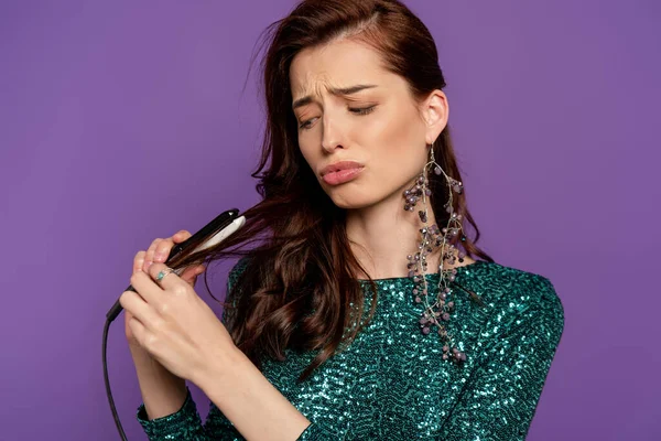 Displeased woman holding small hair straightener near shiny hair isolated on purple — Stock Photo