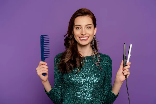 Joven mujer alegre sosteniendo plancha de pelo y peine aislado en púrpura - foto de stock