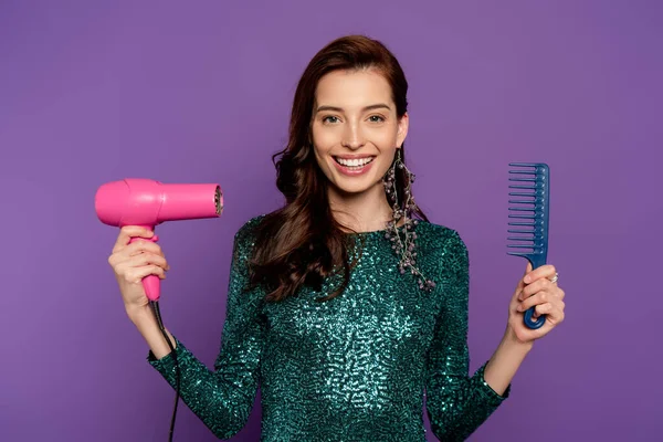 Молодая веселая женщина держит фен и расческа изолированы на фиолетовый — стоковое фото