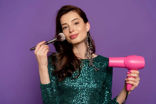 Молода весела жінка тримає фен і косметичний пензлик біля обличчя ізольовано на фіолетовому — стокове фото