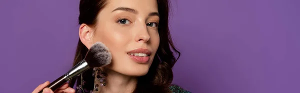 Horizontal concept of young woman holding cosmetic brush while applying blush isolated on purple — Stock Photo