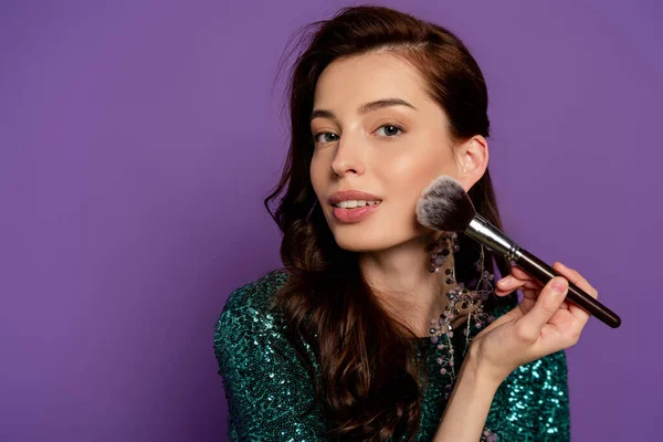 Young woman holding cosmetic brush while applying blush isolated on purple — Stock Photo