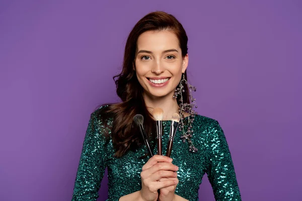 Happy woman holding cosmetic brushes isolated on purple — Stock Photo