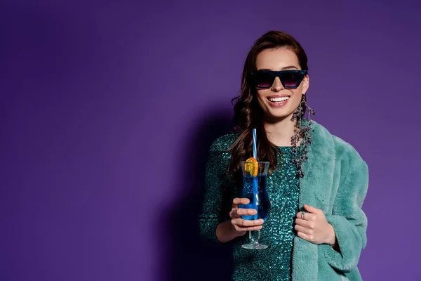 Glückliche Frau mit Sonnenbrille lächelt und hält Stroh in der Nähe von Glas mit Cocktail auf lila — Stockfoto