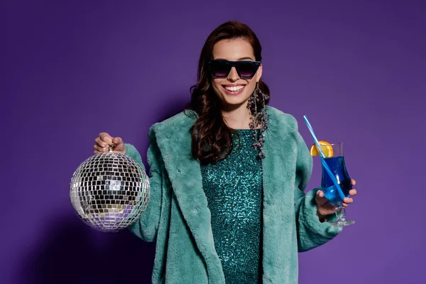 Happy woman in sunglasses smiling while holding glass with cocktail and disco ball on purple — Stock Photo
