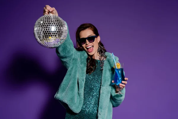 Щаслива жінка в сонцезахисних окулярах тримає скло з коктейлем і диско-кулькою на фіолетовому — стокове фото