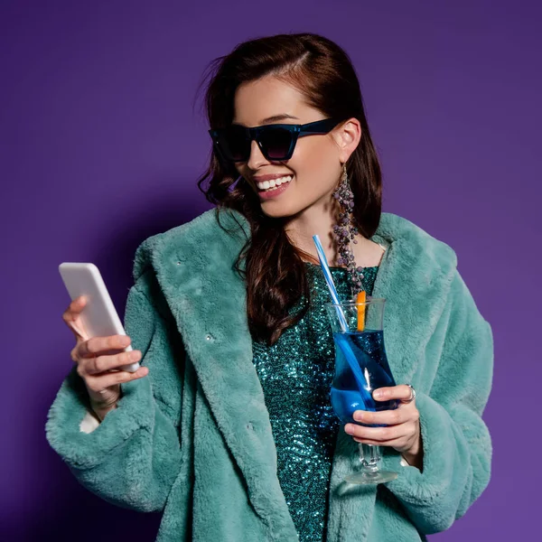 Femme gaie dans des lunettes de soleil tenant verre avec cocktail et smartphone sur violet — Photo de stock