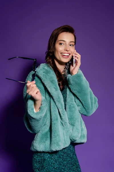 Cheerful girl talking on smartphone and holding sunglasses on purple — Stock Photo