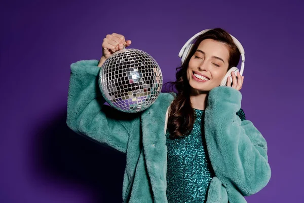 Mulher alegre tocando fones de ouvido e segurando bola de discoteca em roxo — Fotografia de Stock