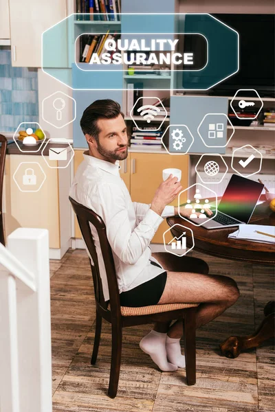 Enfoque selectivo del hombre guapo en camisa y bragas usando el ordenador portátil y sosteniendo la taza en la sala de estar, ilustración de garantía de calidad - foto de stock