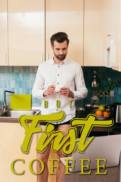 Выборочный фокус красивого мужчины в трусиках и рубашке, смотрящего на ноутбук, когда он пьет кофе на кухне, но первая иллюстрация кофе — стоковое фото