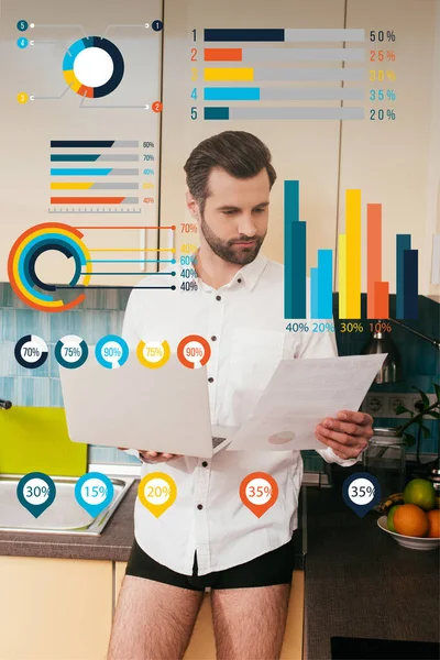 Hombre guapo en camisa y bragas trabajando con documento mientras sostiene el ordenador portátil en la cocina, gráficos y gráficos ilustración - foto de stock