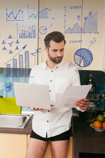 Hombre guapo en camisa y bragas trabajando con documento mientras sostiene el ordenador portátil en la cocina, gráficos y gráficos ilustración - foto de stock