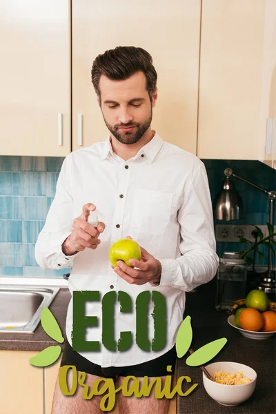 Чоловік у сорочці та трусиках тримає яблуко та дезінфікуючий засіб для рук біля зернових на кухні, екологічно чиста ілюстрація — стокове фото