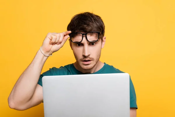 Jovem chocado tocando óculos ao usar laptop no amarelo — Fotografia de Stock