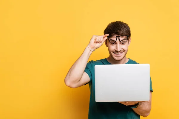 Jovem feliz tocando óculos enquanto usa laptop no amarelo — Fotografia de Stock