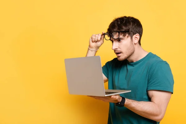 Jovem chocado tocando óculos enquanto olha para laptop no amarelo — Fotografia de Stock