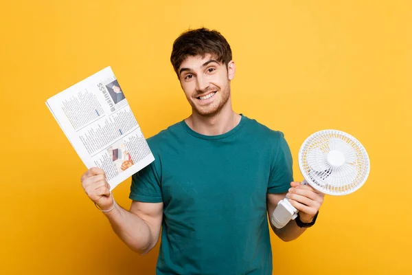 Glücklicher junger Mann hält Zeitung und elektrischen Ventilator auf gelb — Stockfoto