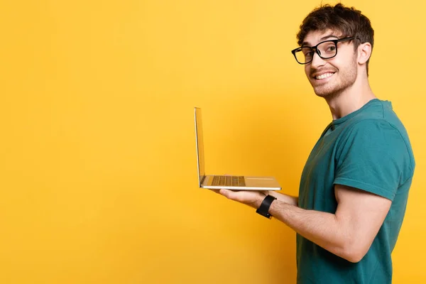 Smiling young man looking at camera while holding laptop on yellow — Stock Photo