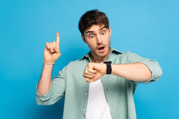 Shocked young man showing idea gesture while looking at wristwatch on blue — Stock Photo