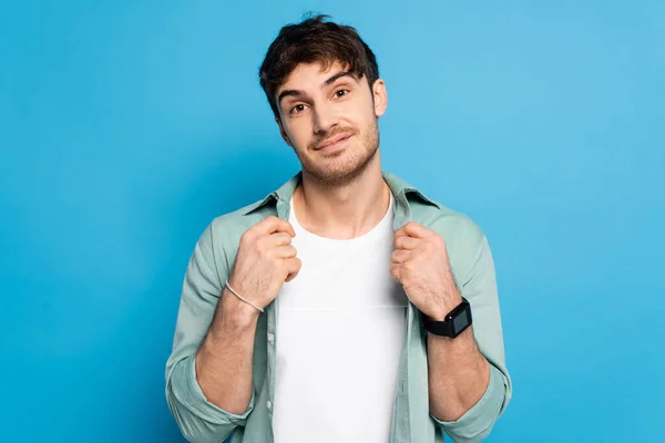 Handsome young man touching shirt while smiling at camera on blue — Stock Photo