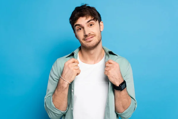 Jovem bonito tocando camisa enquanto sorrindo para a câmera em azul — Fotografia de Stock