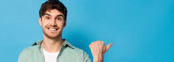 Horizontal image of happy young man pointing with thumb on blue — Stock Photo