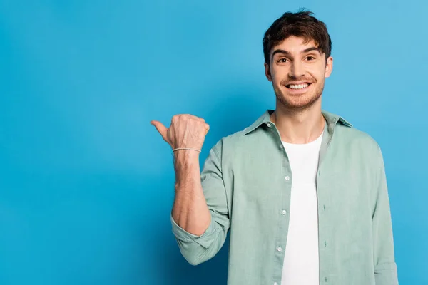 Happy young man pointing with thumb while looking at camera on blue — Stock Photo