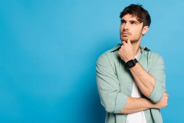 Thoughtful young man looking away and touching chin on blue — Stock Photo