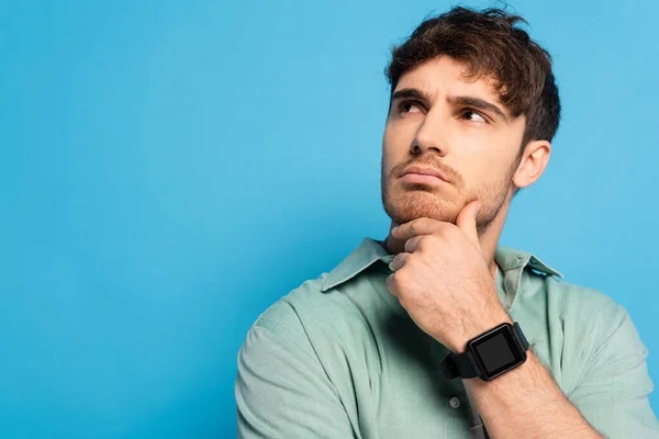 Pensativo joven mirando hacia otro lado mientras toca la barbilla en azul - foto de stock