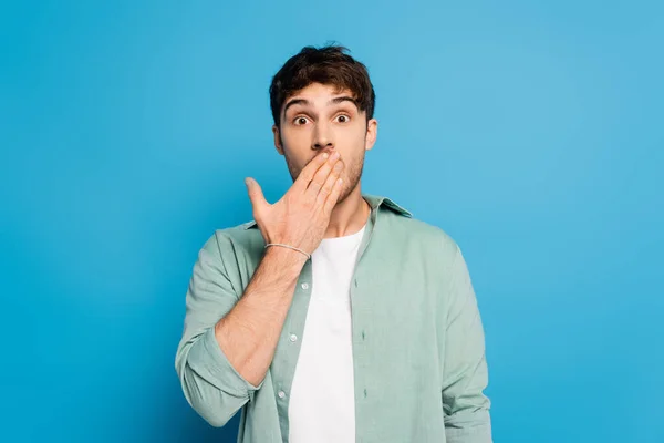 Шокований молодий чоловік прикриває рот рукою на синьому — стокове фото