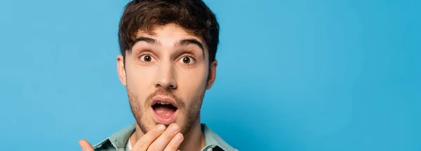 Панорамный снимок шокированного молодого человека, держащего руку у открытого рта, изолированного на голубом — стоковое фото