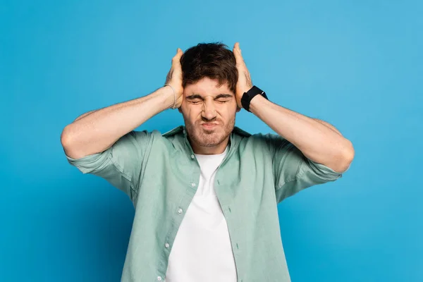 Jovem estressado tocando a cabeça enquanto sofre de dor de cabeça no azul — Fotografia de Stock