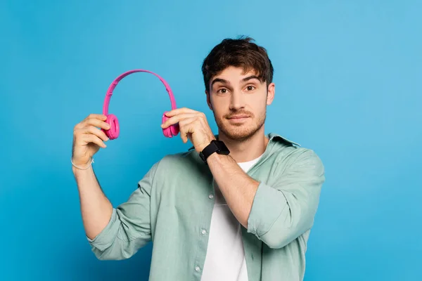 Sonriente joven sosteniendo auriculares inalámbricos mientras mira la cámara en azul - foto de stock