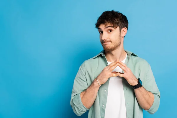 Skeptischer junger Mann steht mit gefalteten Fingern auf blauem Grund — Stockfoto