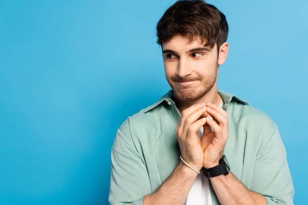 Sly young man looking away while standing with joined fingers on blue — Stock Photo