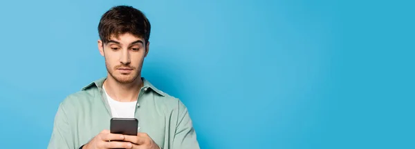 Immagine orizzontale di un giovane uomo serio che chatta su smartphone su blu — Foto stock