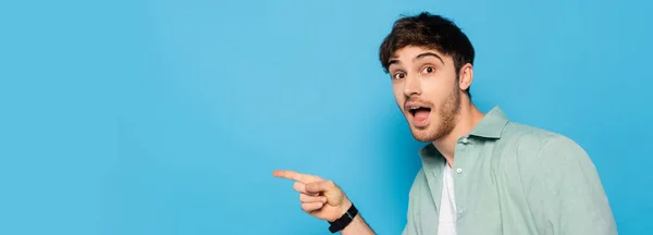 Horizontal image of excited young man pointing with finger and looking at camera on blue — Stock Photo