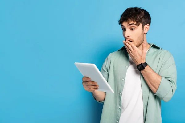 Scioccato giovane uomo che copre la bocca con la mano durante l'utilizzo di tablet digitale su blu — Foto stock