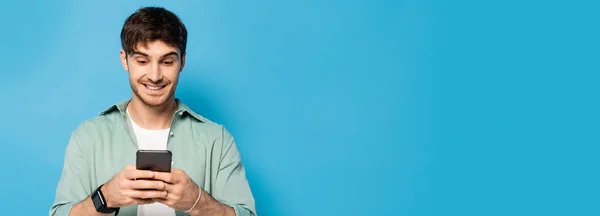 Horizontales Bild eines fröhlichen jungen Mannes, der auf einem Smartphone auf blau chattet — Stockfoto