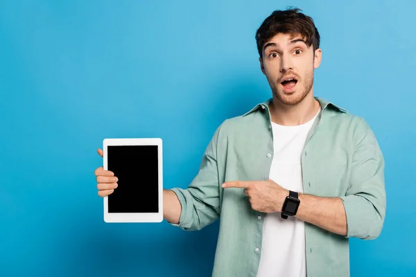 Шокований молодий чоловік вказує на цифровий планшет з порожнім екраном на синьому — стокове фото