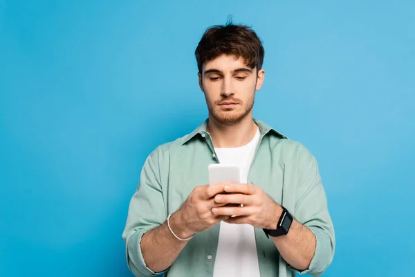 Serious young man chatting on smartphone isolated on blue — Stock Photo
