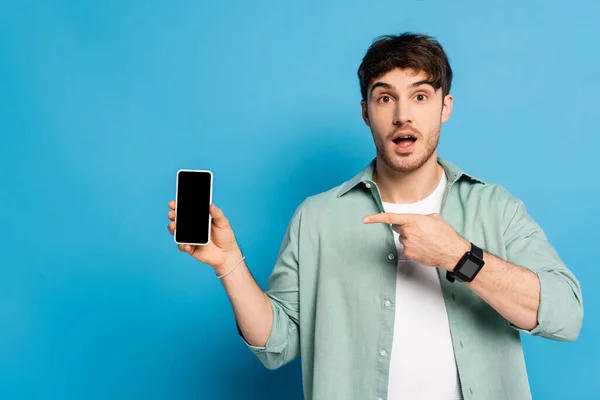 Здивований молодий чоловік вказує на смартфон з порожнім екраном на синьому — стокове фото
