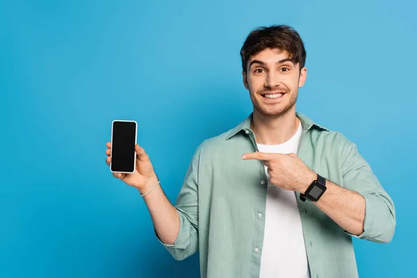 Glücklicher junger Mann zeigt auf Smartphone mit leerem Bildschirm auf blau — Stockfoto