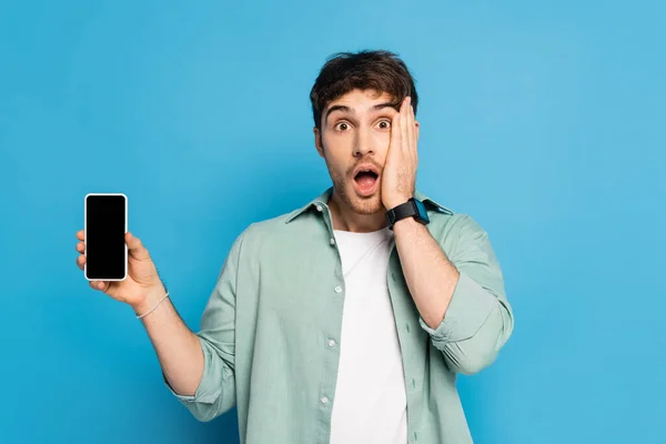 Conmocionado joven tocando la cara y mostrando el teléfono inteligente con pantalla en blanco en azul — Stock Photo